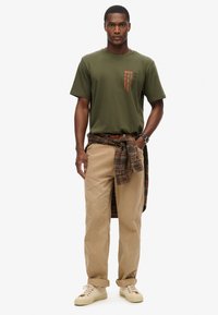 Camiseta verde oliva con texto impreso, pantalones de pana beige, y una camisa de franela a cuadros atada a la cintura. Zapatillas casuales con cordones.