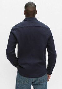 Marineblaues, strukturiertes Hemd mit Button-down-Kragen, langen Ärmeln und lockerer Passform, von hinten gezeigt. Verfügt über zwei Knöpfe an den Manschetten.