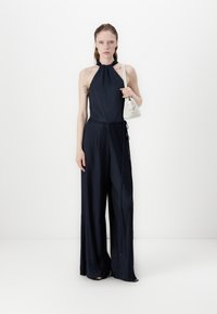 Navy pleated jumpsuit with halter neck design, wide legs, and drawstring waist. Paired with a small white handbag.