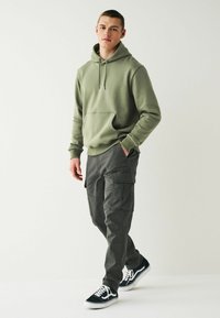 Olivgrüner Kapuzenpullover mit einer Fronttasche, kombiniert mit dunkelgrauen Cargo-Hosen mit Seitentaschen und schwarzen Sneakers mit weißen Schnürsenkeln.