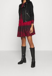 Giacca in suede nera con zip argento sopra un vestito floreale rosso. Stivali neri al ginocchio con tacco spesso. Texture liscia e design a strati.