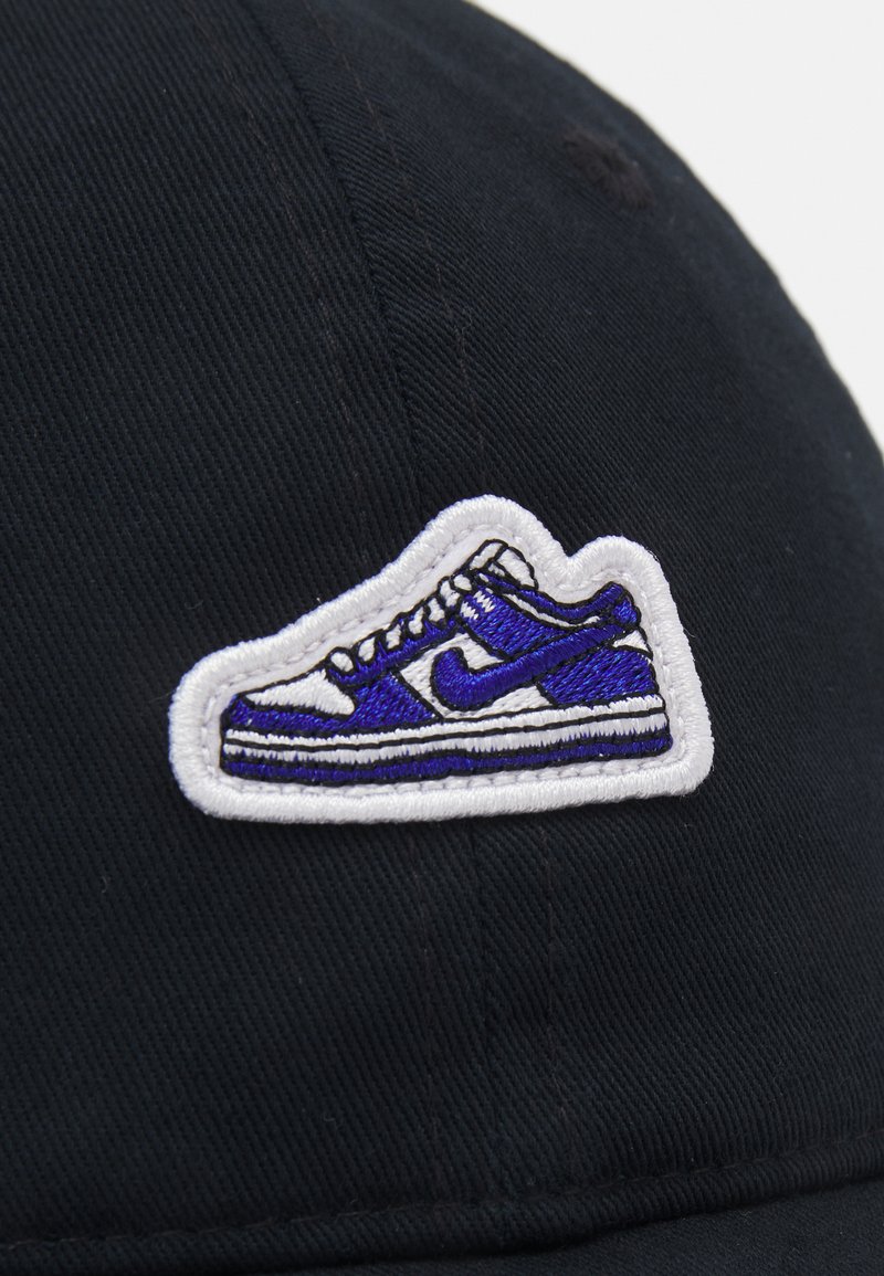 Gorra negra con un diseño de zapatilla en blanco y púrpura bordado en la parte frontal. El material tiene un acabado texturizado con líneas de costura visibles.