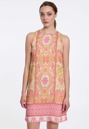 Vrouw draagt een mouwloze jurk met roze en gele bloemenprint en een hoge halslijn, staand tegen een effen witte achtergrond.
