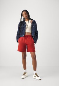 Shorts en coton rouges avec une taille élastique et un cordon de serrage, associés à un crop top beige et à une veste en denim bleue. Baskets beiges et blanches.