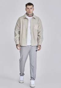 Bež košulja na kopčanje s logotipom, uparen s sivim trenirkama. Košulja ima opušteni kroj, dok hlače imaju suženi dizajn.