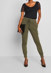 Vrouw in top met zwarte kanten mouwen, olijfgroene cargo joggers, zwarte hoge hakken, met een zwarte clutch, poserend tegen een effen grijze achtergrond.