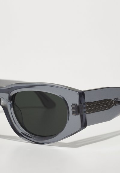 Lunettes de soleil avec une monture grise translucide, des verres foncés et des accents noirs texturés sur les côtés. Design moderne et angulaire avec des branches épaisses.