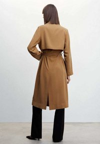 Brown belted trench coat with a collar, long sleeves, and back vent, paired with black wide-leg trousers. Smooth fabric with minimal seams.