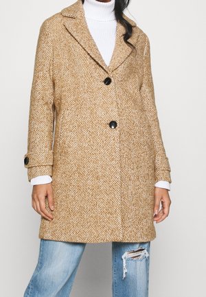 Kvinde iført en tanfarvet frakke med sildebensmønster og sorte knapper over en hvid rullekravebluse og slidte blå jeans.