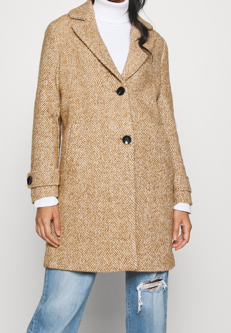 Femme portant un manteau beige à motif chevrons avec des boutons noirs, par-dessus un pull à col roulé blanc et un jean bleu déchiré.