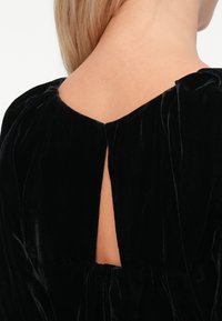 Rochie din catifea neagră cu un decolteu adânc la spate, având un detaliu tip ochi și talie adunată. Textura netedă și luciul delicat adaugă interes vizual.