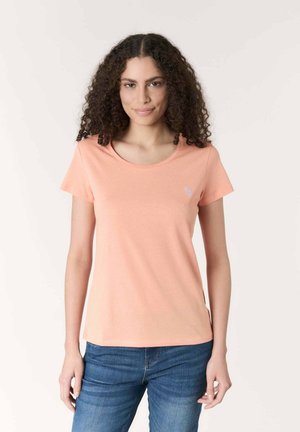 Donna con capelli ricci che indossa una maglietta pesca a maniche corte e jeans blu, in piedi davanti a uno sfondo chiaro e semplice.