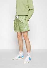 Zelené nylonové šortky s elastickým pasem a šňůrkou, s bílým logem Nike. V páru s bílými a modrými vysokými teniskami.