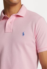 Homme portant un polo rose clair avec un logo brodé bleu de cheval et cavalier sur la poitrine gauche, visible du cou jusqu'à la moitié du torse.