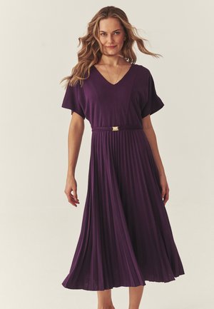 Kvinde med løst, bølget hår iført en bæltefast, kortærmet, plisseret midi-kjole i dyb lilla, stående foran en ensfarvet lyse baggrund.