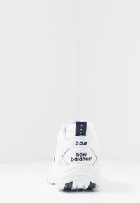 Bílá atletická bota s tmavě modrými akcenty, vybavená polstrovaným límcem, texturovaným designem a značkou "NEW BALANCE" na patě a boku.