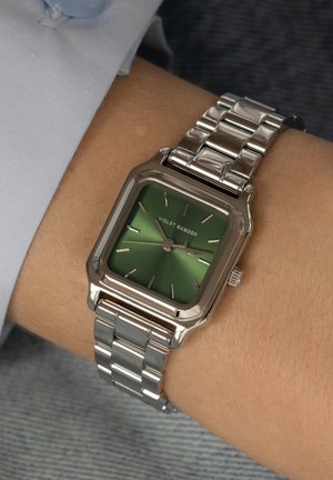 Silberne rechteckige Uhr mit grünem Zifferblatt, silbernen Stundenmarkern und einem Armband aus Edelstahl, getragen an einem Handgelenk vor einem hellen Hintergrund.