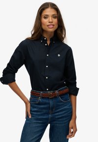 Camisa de botones azul marino con mangas largas, botones blancos y logo bordado, combinada con jeans azules y un cinturón de cuero trenzado marrón.