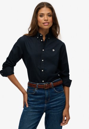 Camisa de botones azul marino con mangas largas, botones blancos y logo bordado, combinada con jeans azules y un cinturón de cuero trenzado marrón.