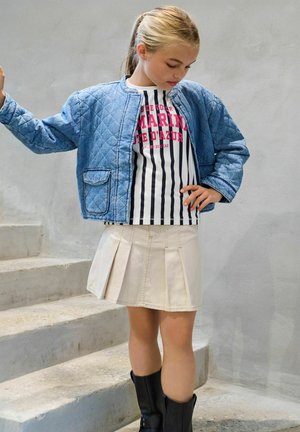 Ung jente i denim quiltet jakke, stripete skjorte, beige plissert skjørt og svarte støvler som står på betongtrapper.