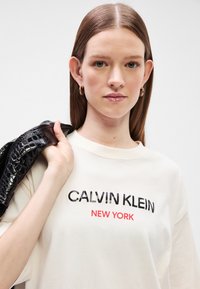 Κρεμ μπλουζίτσα από βαμβάκι με μαύρο και κόκκινο κείμενο "CALVIN KLEIN NEW YORK", άνετη εφαρμογή, κρατώντας μια γυαλιστερή μαύρη τσάντα ώμου.