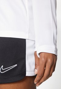 Bijela sportska majica dugačkih rukava s crnim obrubom i mrežastom teksturom, u kombinaciji s crnim šortsevima koji na rubu imaju bijeli Nike logo.