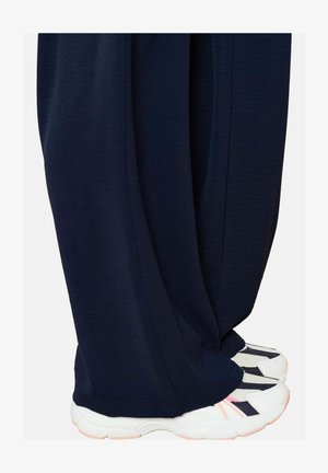 Pantaloni a gamba larga color navy con una finitura testurizzata, abbinati a sneaker bianche con accenti navy e rosa, che mostrano un look sovrapposto e rilassato.