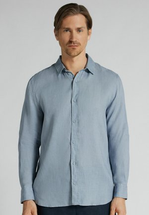 Man met lichtbruin haar die een lichtblauw overhemd met knopen en lange mouwen draagt, staand tegen een effen grijze achtergrond.