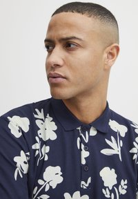 Marineblå polo med hvitt blomsterpattern, med krage og tre knappestolper. Teksturert stoff, tettsittende silhuett.