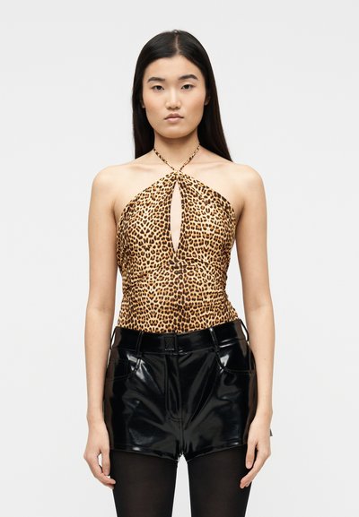 Jeune femme portant un haut dos-nu à imprimé léopard avec une découpe avant, un short noir brillant et des collants noirs, debout devant un fond blanc.
