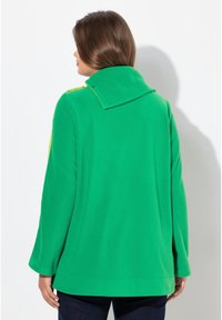 Abrigo verde con cuello alto, confeccionado en una tela suave. Presenta mangas largas y acentos amarillos a lo largo del cuello. Se muestra vista trasera.