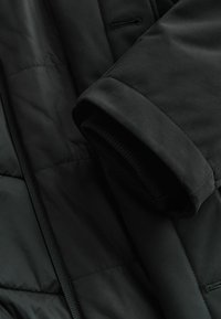 Chaqueta aislante negra con un exterior suave, puños acanalados y un diseño elegante. Presenta una textura acolchada y bolsillos ocultos.