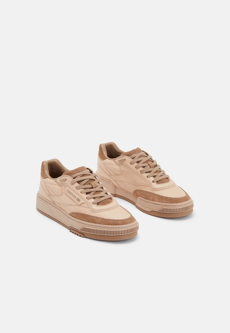 Beige leather and suede sneakers with flat laces, padded collar, and rubber sole featuring a textured grip pattern. Brand logo on side.