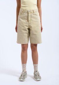 Pantalones cortos de mezclilla beige con cintura alta, cinco bolsillos y cierre de botón; combinados con zapatillas grises y calcetines ligeros y texturizados.
