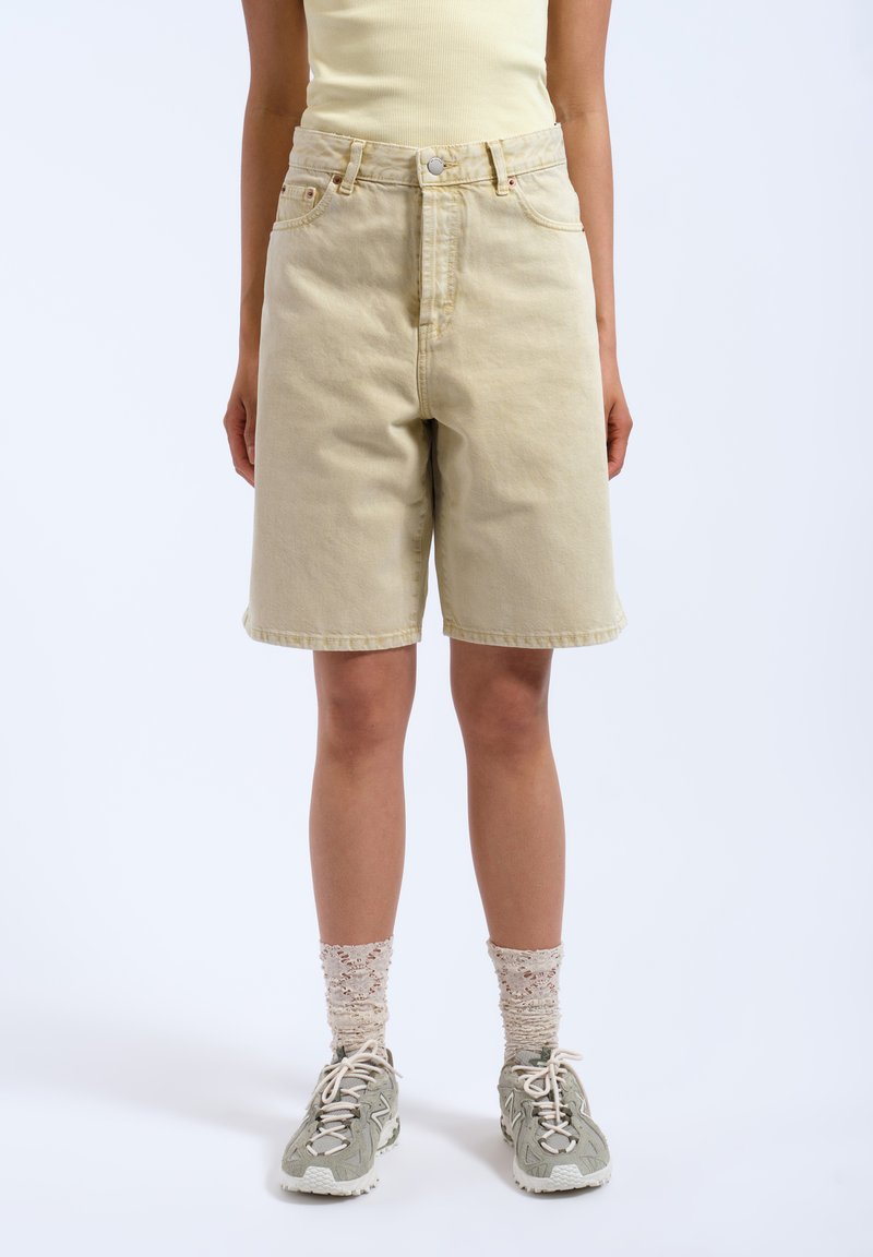 Pantalones cortos de mezclilla beige con cintura alta, cinco bolsillos y cierre de botón; combinados con zapatillas grises y calcetines ligeros y texturizados.
