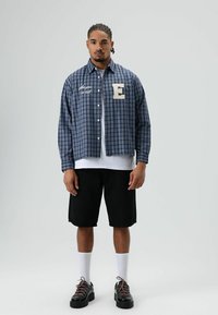 Blauw-wit geruit flanellen overhemd met knoopsluiting, geborduurde "E", gedragen over een wit T-shirt, gecombineerd met zwarte korte broek en sneakers.