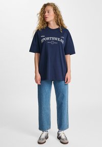 Mörkblå oversized t-shirt med texten "SPORTSWEAR", parat med ljusblå raka jeans och grå sneakers med vita snören.
