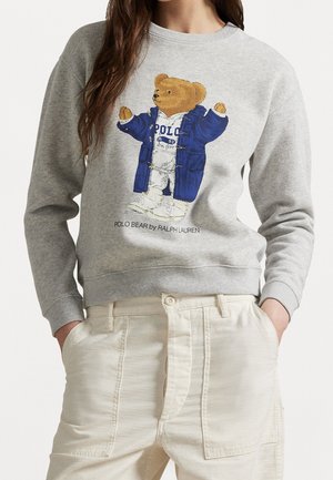 Femme portant un sweat-shirt gris avec un dessin d'ours en peluche en manteau bleu et un pantalon crème, les mains dans les poches.