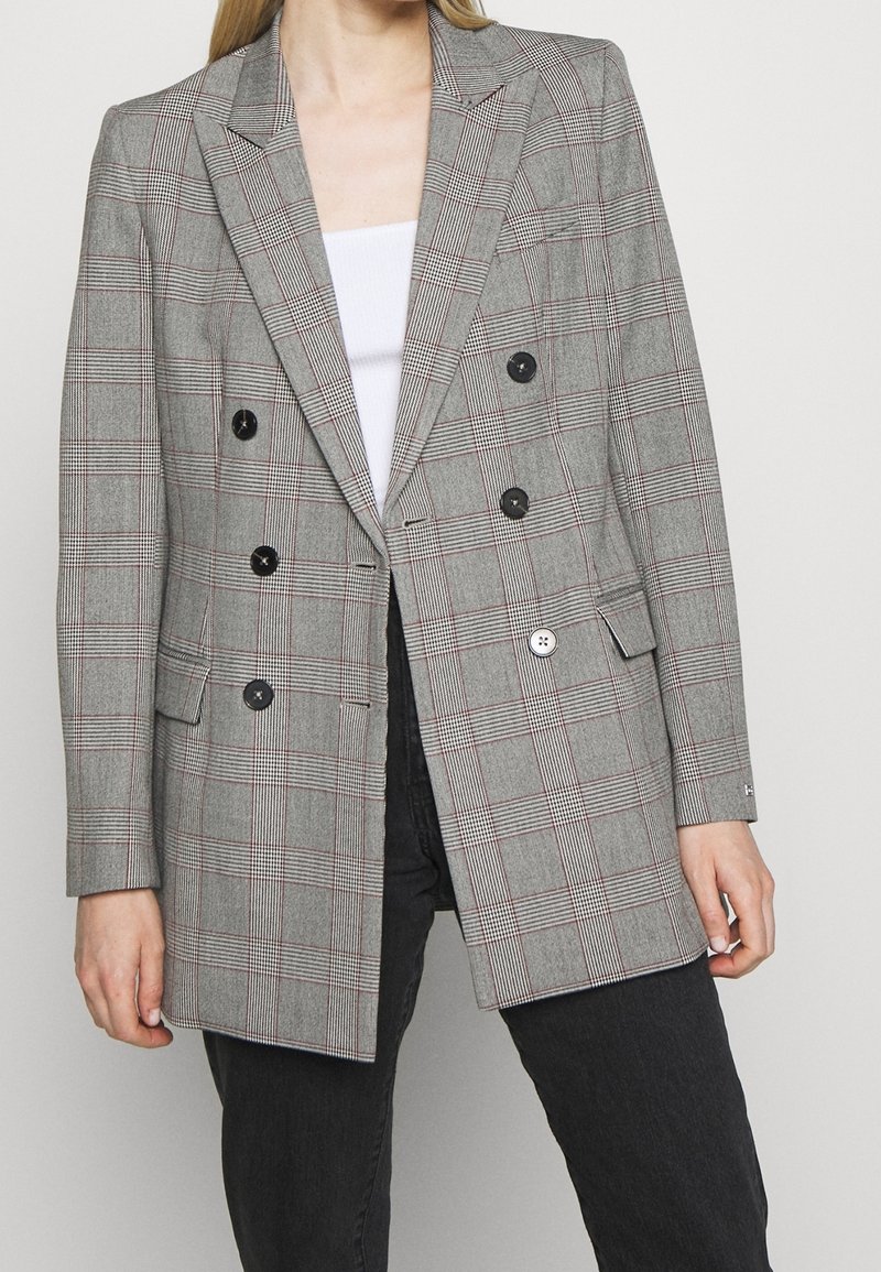 Blazer de cuadros gris de doble botonadura con botones negros, solapas con muescas, bolsillos laterales y un corte ajustado. Tejido texturizado con acentos rojos.