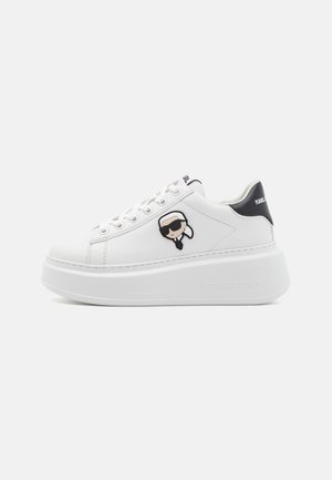 Sneaker de cuero blanco con suela de plataforma, puntera redonda, cordones en la parte delantera y un logo de dibujos animados en el lateral. Acento negro en el talón con el nombre de la marca.