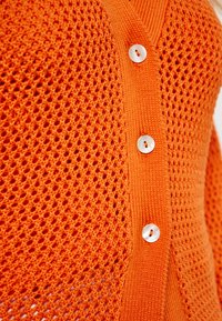Orange stickad kofta med en strukturerad öppen väv, V-ringning och fyra vita knappar fram.