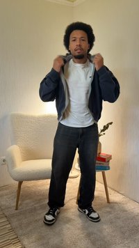 Granatowa kurtka na zamek błyskawiczny noszona na białej koszulce, do tego ciemne dżinsy i czarno-białe trampki. W tle jasne, lekkie fotel i dekoracje.