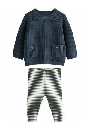 Dunkelblauer Strickpullover mit zwei aufgenähten Taschen und Holzknöpfen, kombiniert mit elastischen Hosen in Creme- und Marineblau mit Streifen.