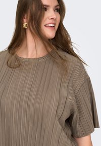 Χαμογελαστή γυναίκα με μακριά καστανά μαλλιά, που φορά μεγάλα χρυσά σκουλαρίκια και ένα πιετεντό τούπα κοντομάνικο τοπ, κοιτάζει προς τα πλάγια.