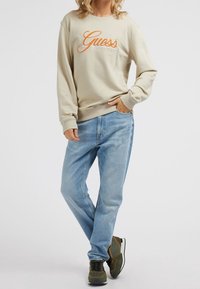 Sweat-shirt beige avec logo "Guess" orange, associé à un jean bleu clair à coupe ample et des baskets vert-noir avec des accents orange.