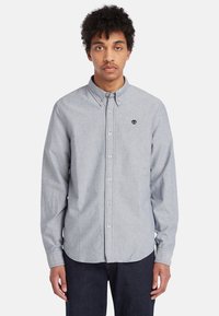 Camisa de botones en gris claro con cuello, corte clásico y textura sutil. Presenta un pequeño logo en el lado izquierdo del pecho y botones blancos.