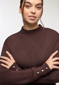 Maglione a collo alto lavorato a maglia marrone con maniche lunghe decorate da accenti di cristallo sui polsini. Texture liscia e vestibilità comoda.