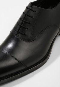 Chaussure Oxford en cuir noir avec une finition lisse, bout arrondi et six œillets pour les lacets. Présente un bout renfort subtil et une silhouette élégante.