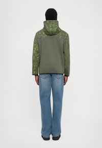 Sweatshirt com capuz em verde escuro, com acentos texturizados e padrões em verde brilhante. Combinado com jeans de perna larga em azul claro.