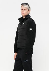 Sort puffer vest med fleeceærmer, lynlås og labels på bryst og ærme. Sammen med sorte joggers med blå tekst.